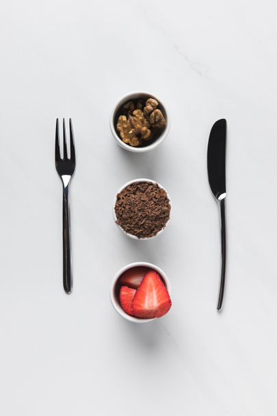Bowls with walnuts, grated chocolate and strawberries between fork and knife, table appointments conception - Photo, Image