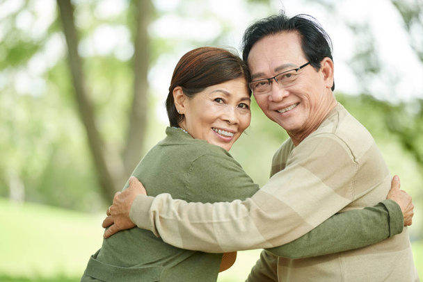 Улыбающаяся старшая азиатская пара обнимается и поворачивается, чтобы посмотреть в камеру
 - Фото, изображение