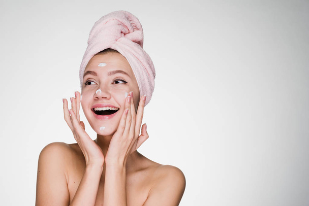 happy young girl with pink towel on head applying moisturizer on face - Foto, Imagem