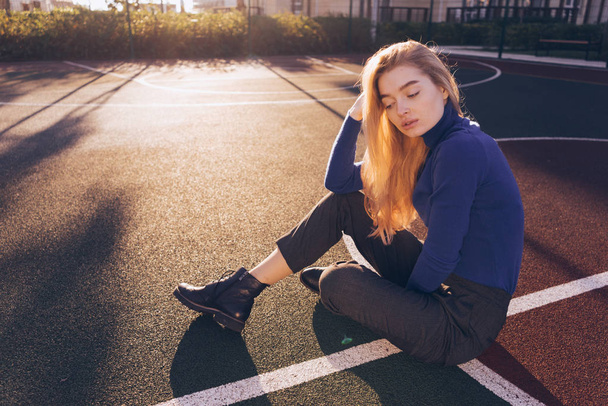 beautiful young long-haired blond girl in blue sweater sits on the sports field in the city, posing, eyes closed - Foto, immagini