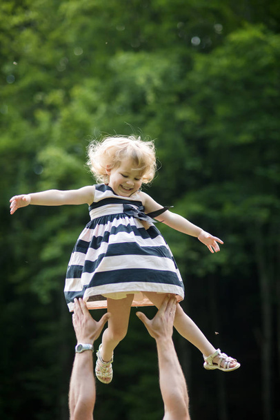 Girl smile flying to father hands on sunny summer day on natural environment. Freedom, future, optimism, support. Happy childhood concept - Фото, изображение