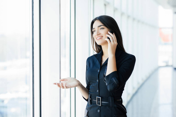 Young businesswoman talking on mobile phone while standing by window in office. Beautiful young female model in bright office. - Foto, imagen
