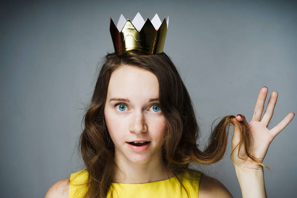 beautiful surprised young girl with a golden crown on her head holds a curl of hair in her hand - Photo, Image