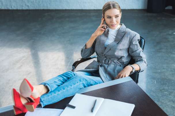 high angle view of businesswoman sitting with legs on table in office and talking by smartphone - Valokuva, kuva