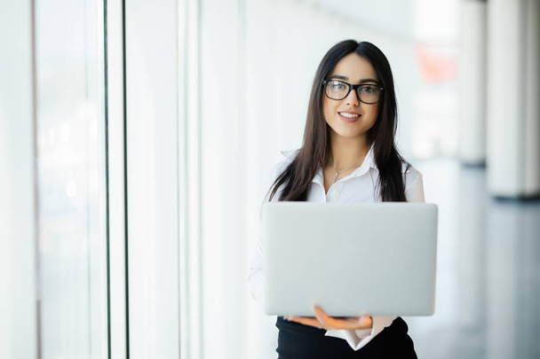 Молодая деловая женщина, работающая в своем роскошном офисе, держит ноутбук, стоящий напротив панорамного окна с видом на деловой район
 - Фото, изображение