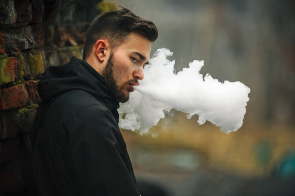 Vape man. Photo of a handsome young white guy with modern haircut in the black clothes vaping and letting off steam from an electronic cigarette near - Photo, image