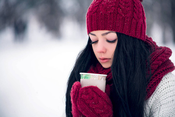 Güzel genç bir kadın beyaz bir kazak ve parlak bir şapka, eşarp ve eldiven bir bardak kahve ile - Fotoğraf, Görsel