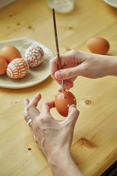 female hands holding brush and egg over table with painted eggs with various white patterns for Easter holiday on plate        - Foto, Imagem