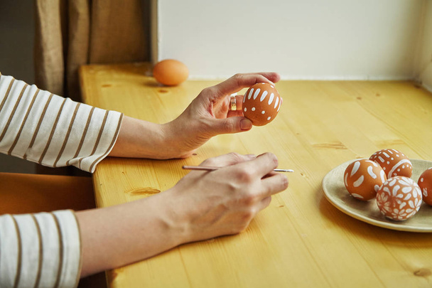 female hands holding brush and egg over table with painted eggs with various white patterns for Easter holiday on plate        - Photo, Image