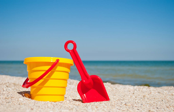 Yellow childrens bucket with red toy toy plastic red scapula on the left against the blue sea and blue sky yellow sand beach sea shore seashells summer vacation sunny summer day, bright sun, baby toys - Photo, Image