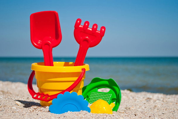 Yellow baby bucket with red handle, plastic red spatula and rake, green sieve, yellow sand form of strawberry, blue plate, blue sea and sky, yellow sand beach seashells seaside summer vacation sunny - Photo, Image