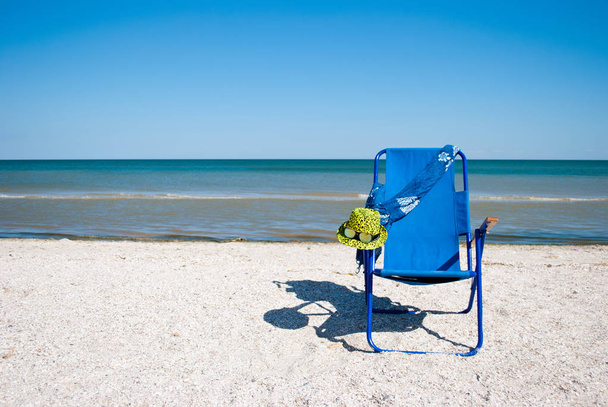 Sininen ranta tuoli ja tyylikäs keltainen hattu aurinkolasit rannalla sininen meri sininen taivas valkoinen pilvet kaunis maisema kesä loma ranta hiekka viikonloppu levätä rannalla
 - Valokuva, kuva