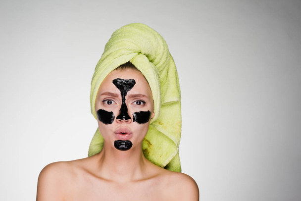 an indignant young girl with a green towel on her head applied a black mask to the problem zones on her face - Foto, Bild