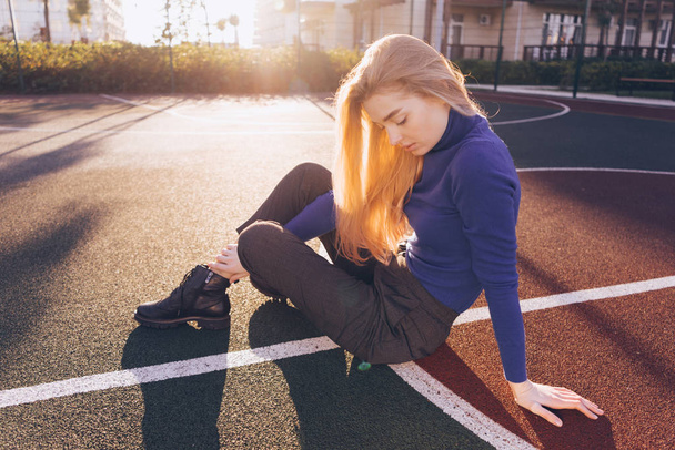 stylish beautiful young blond girl in a blue sweater sits on a sports field in the sun - Photo, image