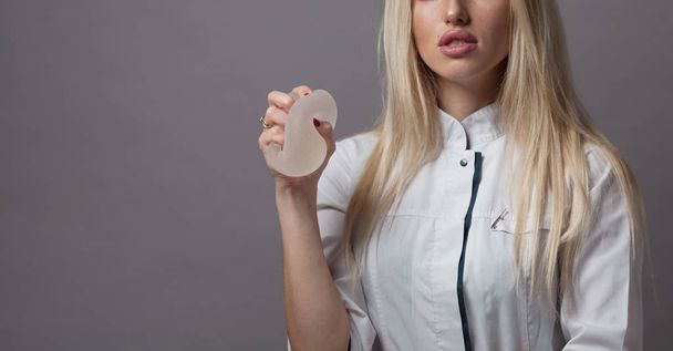 Plastic surgeon woman holding silicon breast implant on grey background. Plastic surgery concept - Фото, изображение