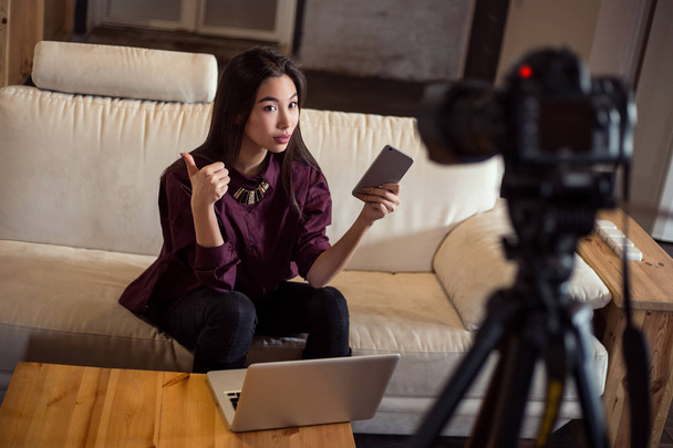 Όμορφη νεαρή κουκλίτσα-κορίτσι vlogger κάθεται στον καναπέ με τα smartphone και laptop και κρατώντας ένα blog χρησιμοποιώντας μια φωτογραφική μηχανή με τρίποδα. - Φωτογραφία, εικόνα
