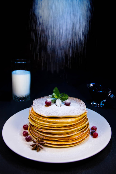 Pannenkoeken op een witte plaat met cranberry puddingen en kaneel besprenkeld met suiker poeder samen met bosbessen jam en een glas melk - Foto, afbeelding