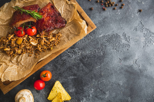 Grilled pork ribs, buckwheat with mushroom and tomatoes on vintage wooden cutting board. Flat lay, top view. Copy space. - Photo, Image
