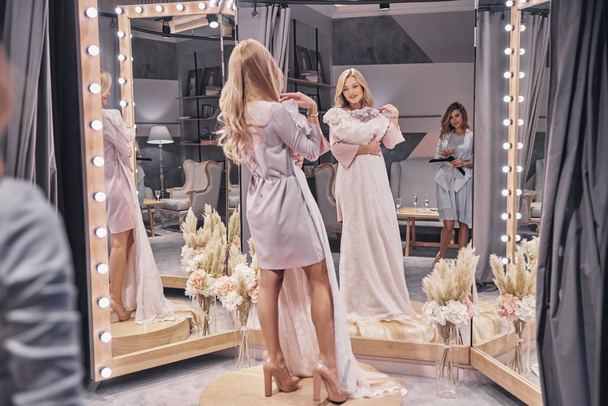 full length of beautiful blonde woman choosing wedding dress in fitting room with bridesmaid - Фото, зображення