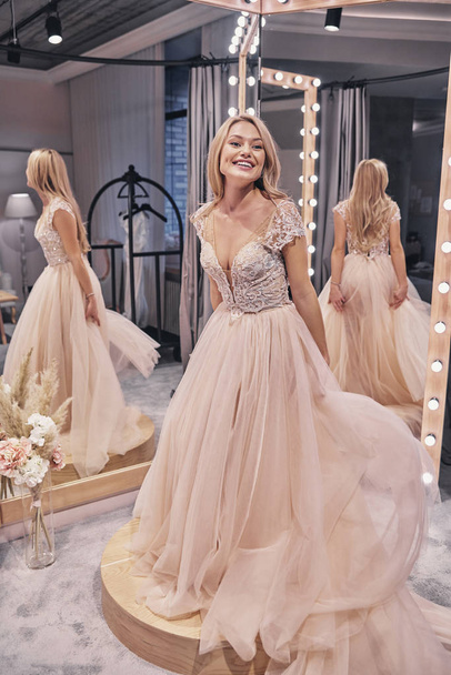 happy woman wearing wedding dress and smiling in front of mirror in bridal shop - Valokuva, kuva