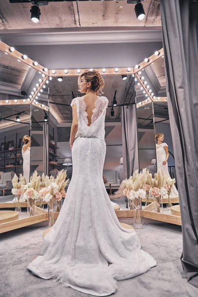Full length of gorgeous young woman wearing amazing wedding dress while standing in front of the mirror in bridal shop - Photo, Image