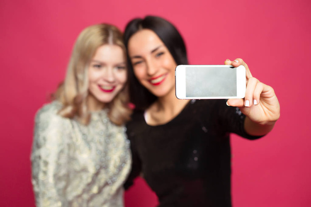 Two cheerful and beautiful smiling girlfriend photographed themselves on the phone or make a selfie on a pink background. - Фото, изображение