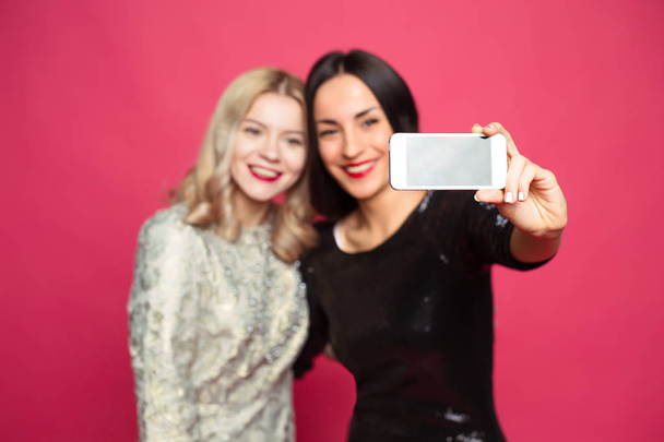 Two cheerful and beautiful smiling girlfriend photographed themselves on the phone or make a selfie on a pink background. - Photo, image
