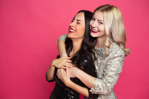 Friends forever. Close up photo of Two happy young beautiful smiling girlfriends in little black dresses posing and having fun on a pink background. - Фото, изображение