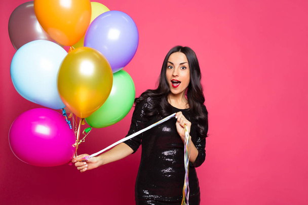 Girl with helium balloons. Beautiful smiling stylish woman in a little black dress with colorful helium balloons on a pink background. - Фото, изображение