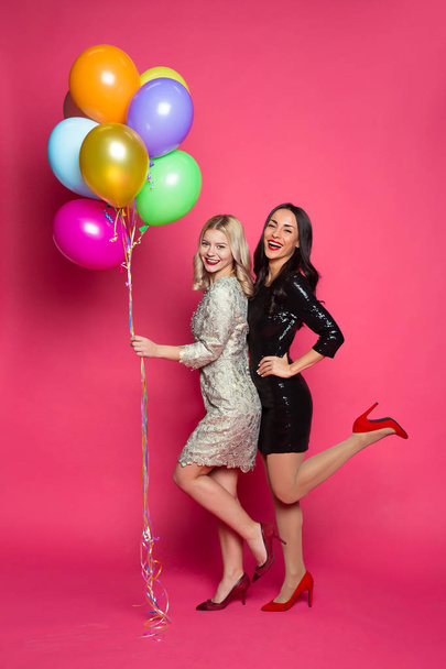 Funny women. Two happy and cheerful beautiful girl friends in beautiful dresses posing and having fun with helium balloons in hands on a pink background. - Fotoğraf, Görsel