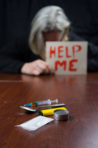 A drug addict at the table holding a sign m asking for help, in front of him a syringe of drugs. - Valokuva, kuva