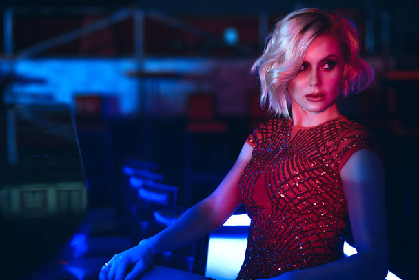 Close up portrait of beautiful glam blond woman with provocative make up wearing red sequin dress sitting at the bar in the night club in colourful neon lights and looking aside - Foto, Imagen