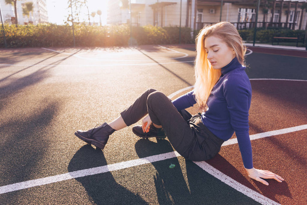 stylish beautiful blond model girl in blue sweater sits on sports field in the sun - Photo, Image