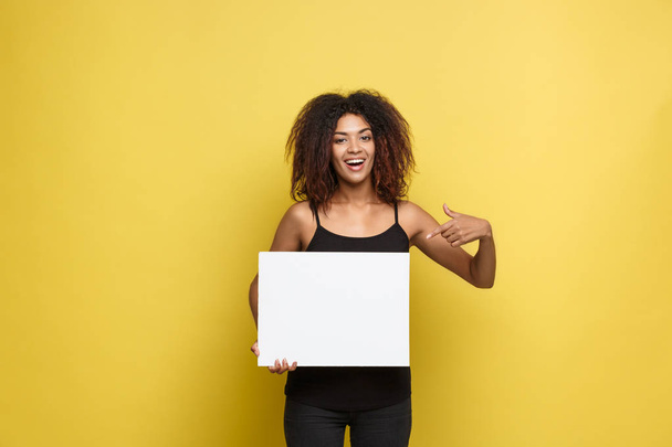 Business Concept - Close up Portrait young beautiful attractive African American pointing finger to plain white blank sign. Yellow Pastel studio Background. Copy space - Fotoğraf, Görsel