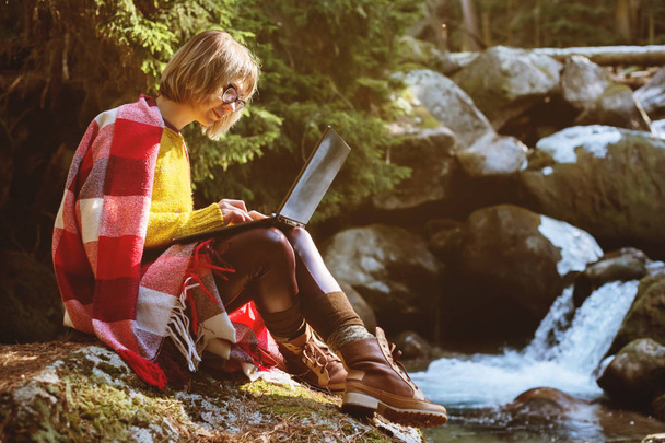 Un ritratto tonico di una ragazza hipster freelance sorridente con gli occhiali vestiti con una coperta con un computer portatile inginocchiato seduto su una roccia in una foresta di conifere accanto a un fiume di montagna tempestoso in caldo
 - Foto, immagini