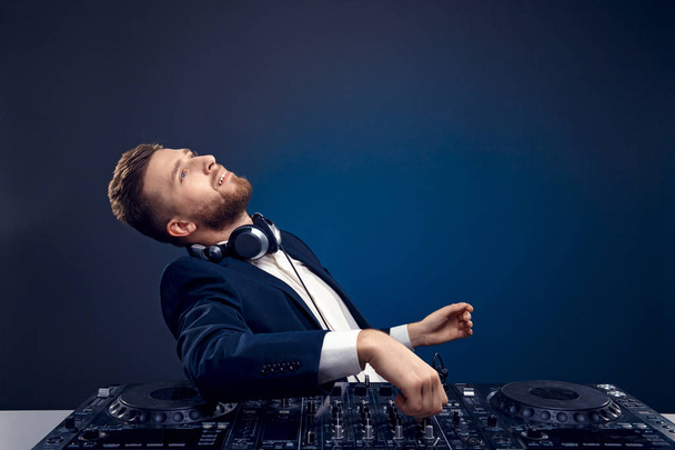 Closeup portrait of confident DJ with stylish hair style and headphones on neck mixing music on mixer while standing isolated on dark colored blue, cyan background - Valokuva, kuva