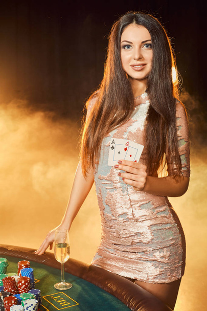 Prachtige jonge vrouw in avondjurk staande in de buurt van pokertafel met glas champagne - Foto, afbeelding