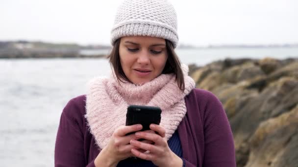 Feliz hermosa mujer en invierno frente a la mensajería del océano con mensajes de texto amante en el teléfono celular - Imágenes, Vídeo