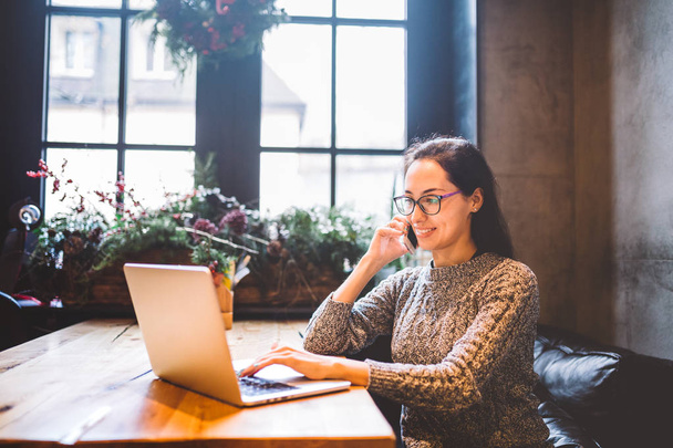 Theme is small business. A young freelance woman working behind a laptop computer in a coffee shop decorated with Christmas decor and talking on the phone. Dressed in a gray sweater and glasses - Photo, Image