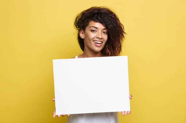 Obchodní koncept - zblízka portrét mladé krásné atraktivní afroamerické usmívající se zobrazeno obyčejné bílé prázdné znamení. Žlutá pastelová studio pozadí. Kopírovat prostor - Fotografie, Obrázek