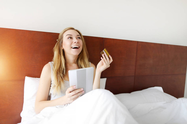 mujer feliz con tarjeta de crédito de débito y tableta puesta en la cama blanca en casa con sol
 - Foto, Imagen