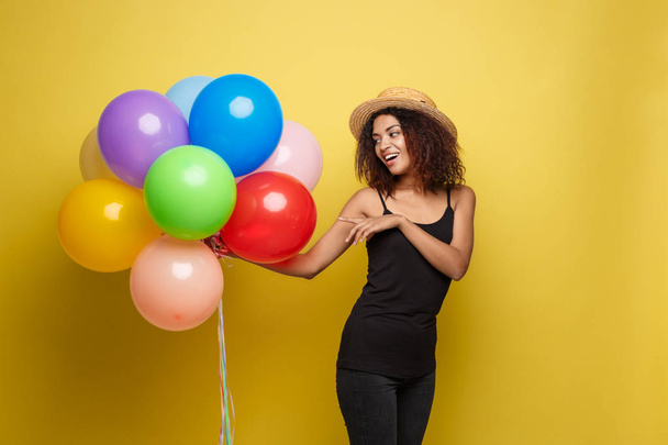 Ünneplés koncepció - portré boldog fiatal gyönyörű afrikai nő fekete póló mosolygós, színes párt ballon közelről. Sárga pasztel stúdió háttér - Fotó, kép