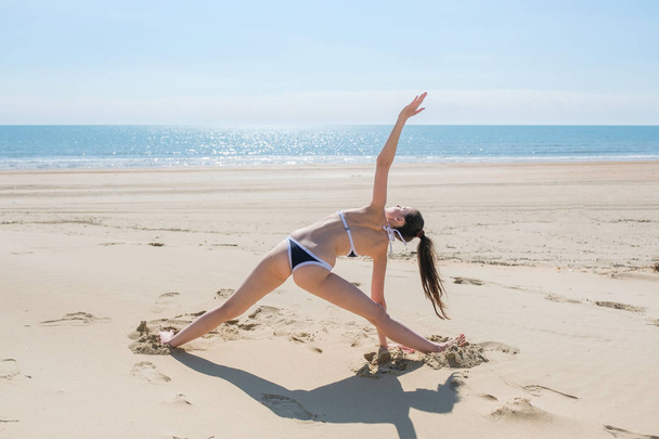 Vrouw in bikini doen yoga staat in de positie van een driehoek op het strand. Terugkijken, weergave. - Foto, afbeelding