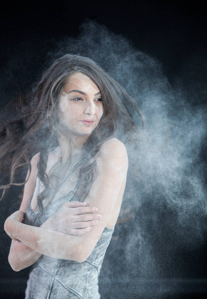 Graceful young beautiful sexy brunette with long hair dancing in cloud of dust. Portrait young beautiful sexy brunette with long hair woman covered with powder on black background - Foto, afbeelding