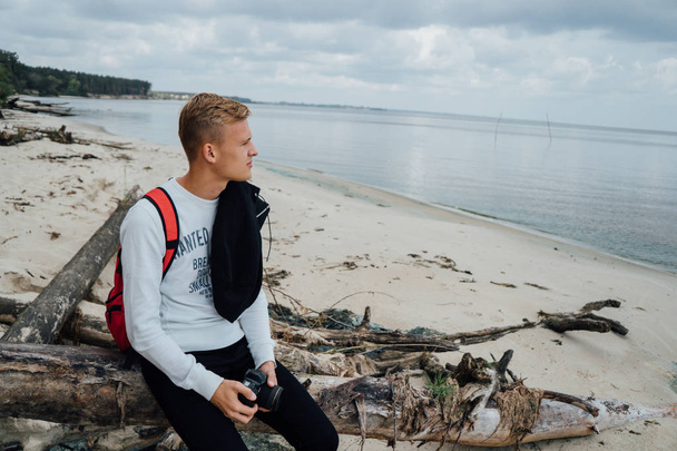 Young blonde man sitting at woods on river shore and looking away with camera in hands - Foto, immagini