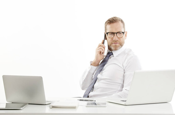 Middle aged male business broker using mobile phone and making call while sitting at desk in front of laptop. Isolated on white background. - Photo, image