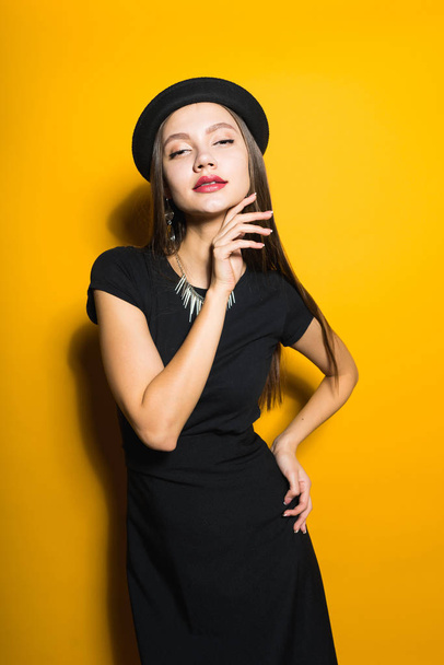 beautiful attractive girl in stylish black hat and dress posing on yellow background - Foto, Imagen