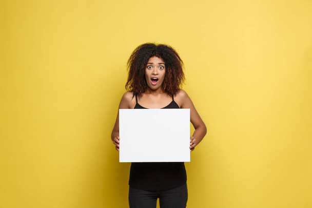 Business Concept - Close up Portrait young beautiful attractive African American shocking expression showing plain white blank sign. Yellow Pastel studio Background. Copy space - Foto, imagen