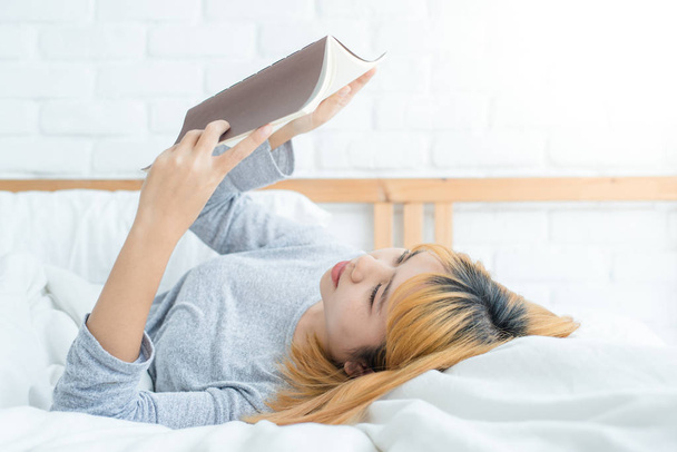 Joven mujer asiática disfrutando tumbada en la cama leyendo libro placer en ropa casual en casa. Concepto relajante
. - Foto, imagen