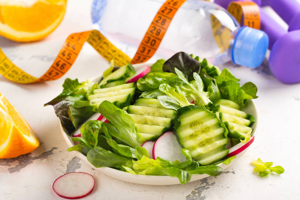 Здоровый вегетарианский салат, гантели и бутылка воды на деревянном столе
 - Фото, изображение